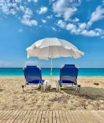 Grafika przedstawiająca dwa niebieskie leżaki stojące na piasku, pomiędzy nimi słoneczny biały parasol, w tle błękitne morze